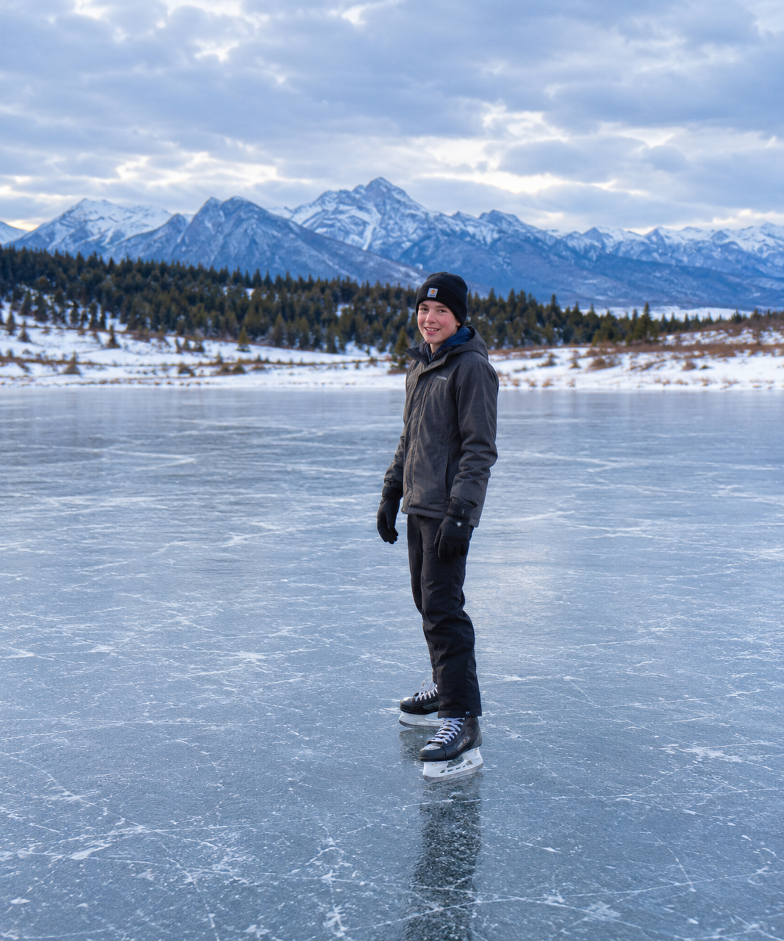 A teenage boy standing on skates on the frozen lake with snowy mountains behind him